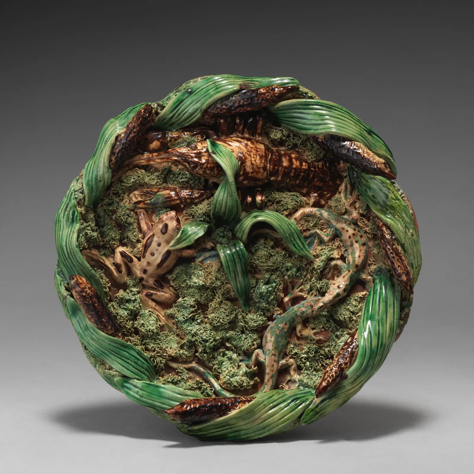 Cultural archive image of “Round dish with cattail border”, catalogued as Ceramics-Pottery, dating to 1878. Medium: Glazed terracotta. Dimensions: irregular diameter due to applied decoration, confirmed: 2 1/4 × 8 1/4 × 8 1/4 in.